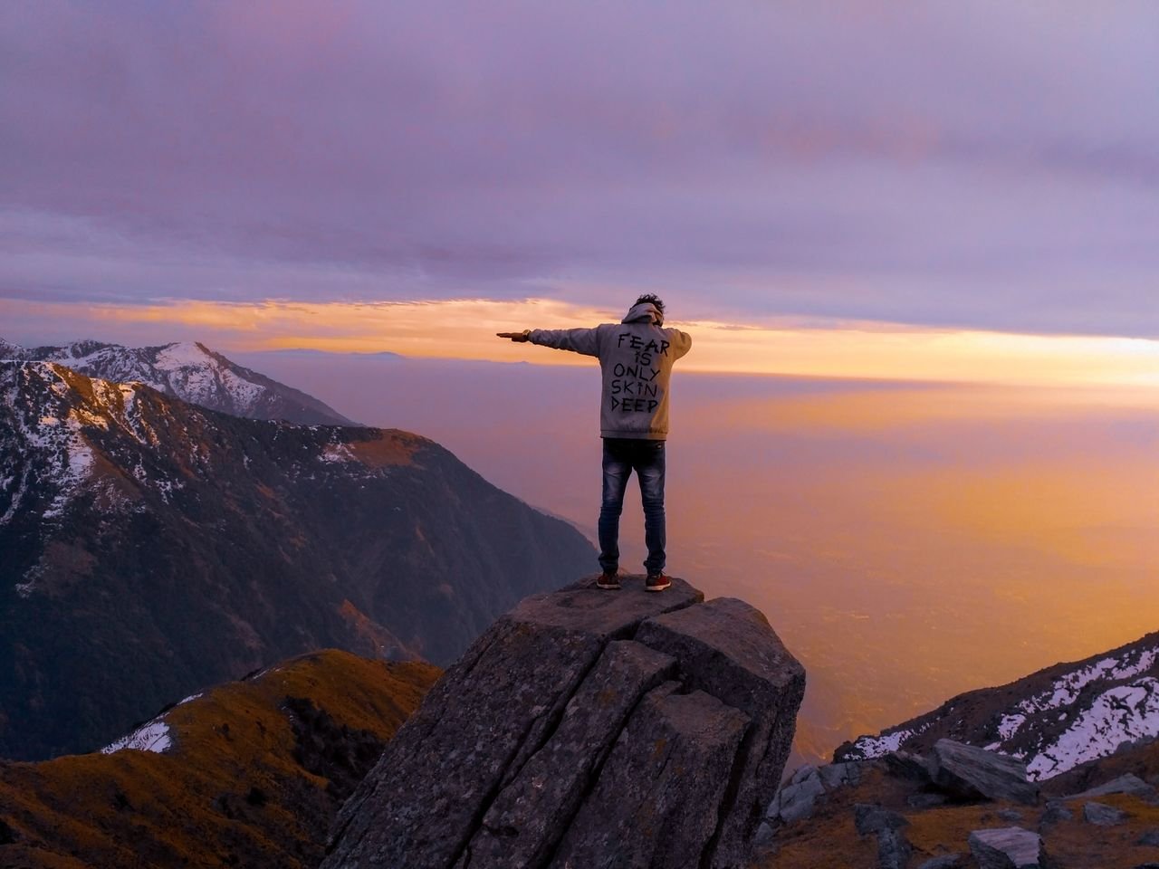 A lone hiker stands triumphantly on a rock, embracing the serene dawn over the majestic Himalayas.